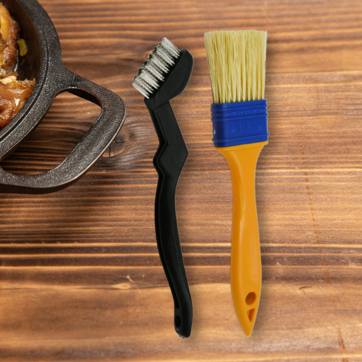 A black grill cleaning brush and a yellow and blue basting brush stand side-by-side on a rustic wooden surface.
