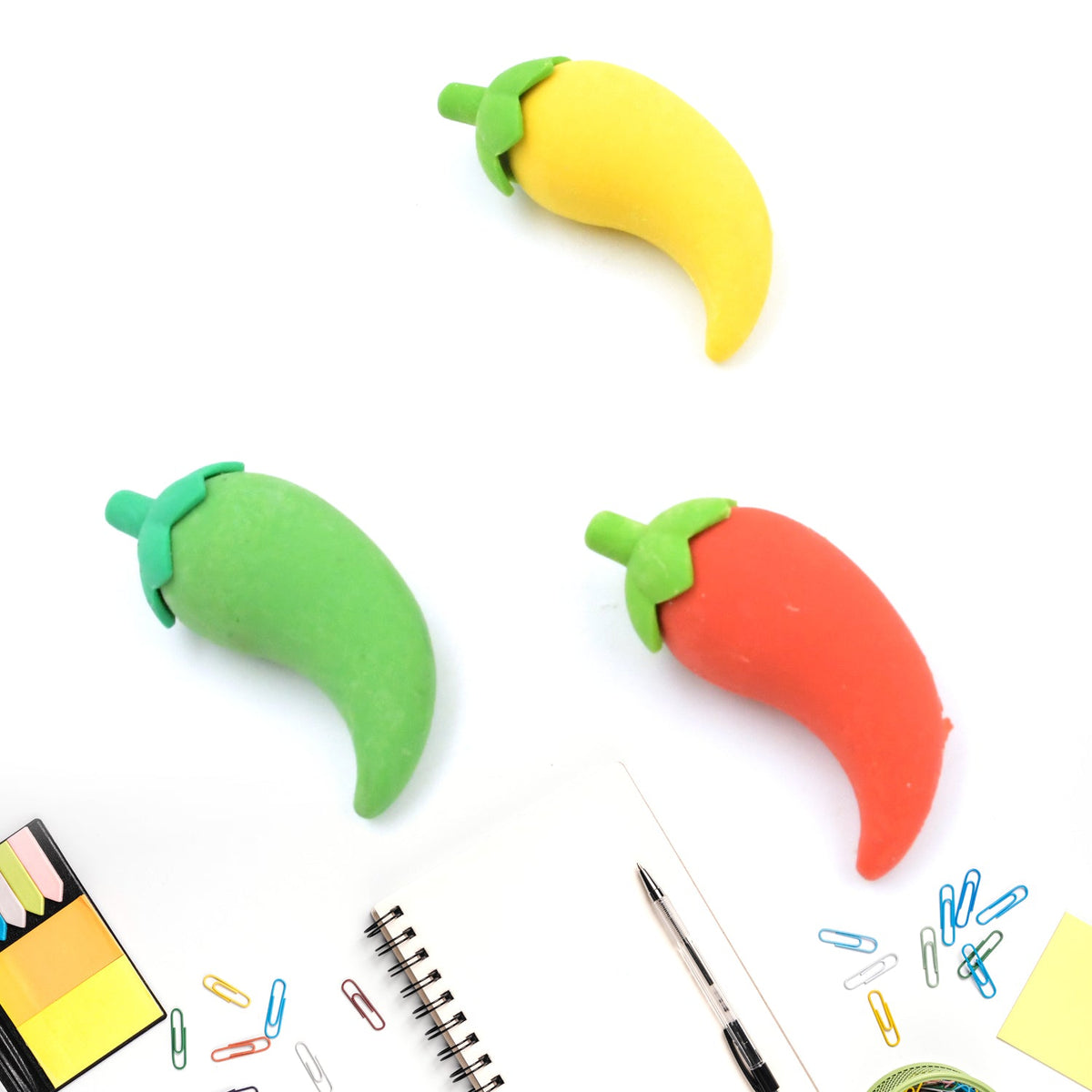 Three chili pepper erasers in red, yellow, and green, arranged on a white surface with a notebook, pen, and paper clips.