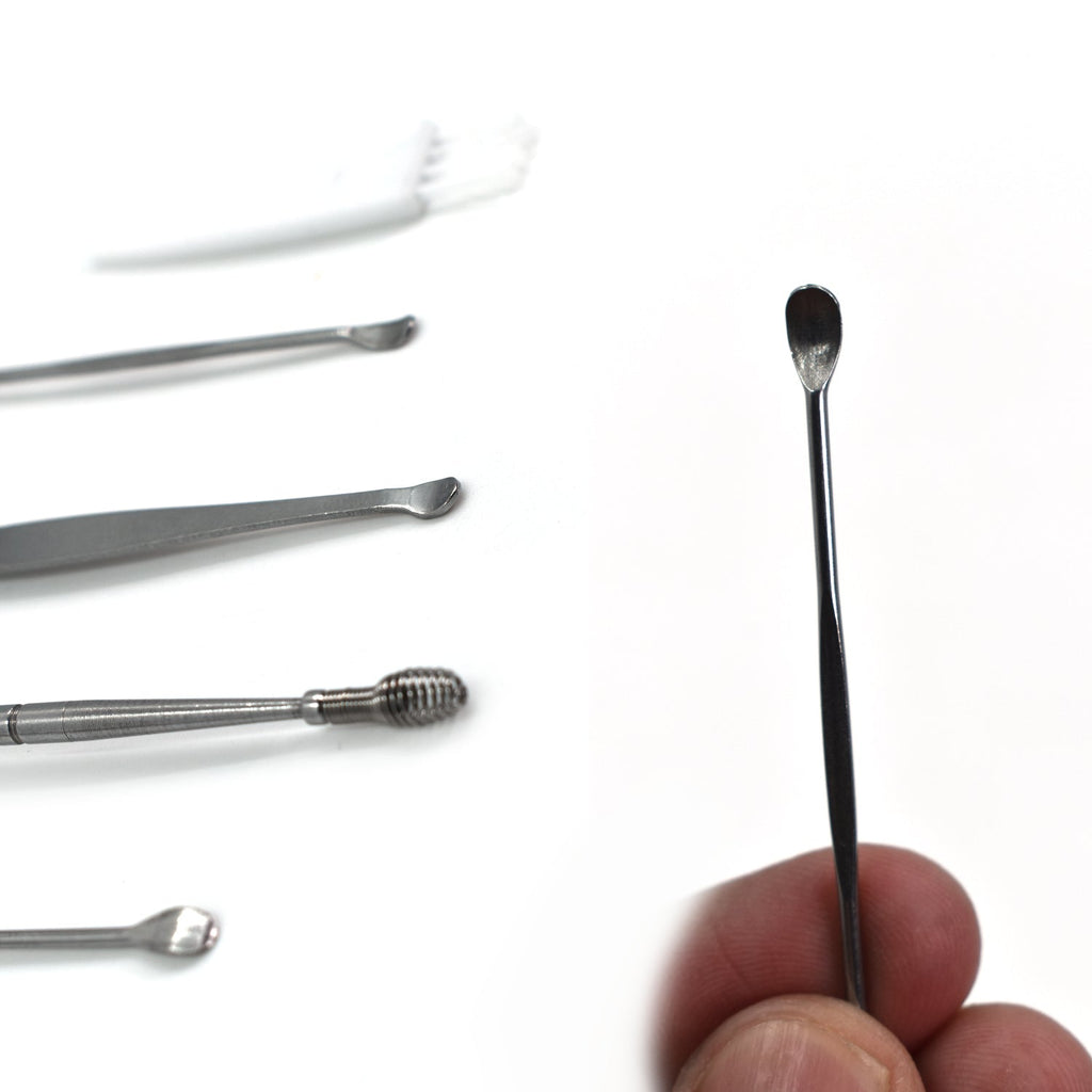 A hand holds one silver ear spoon tool, with other tools and a white brush blurred in the background.