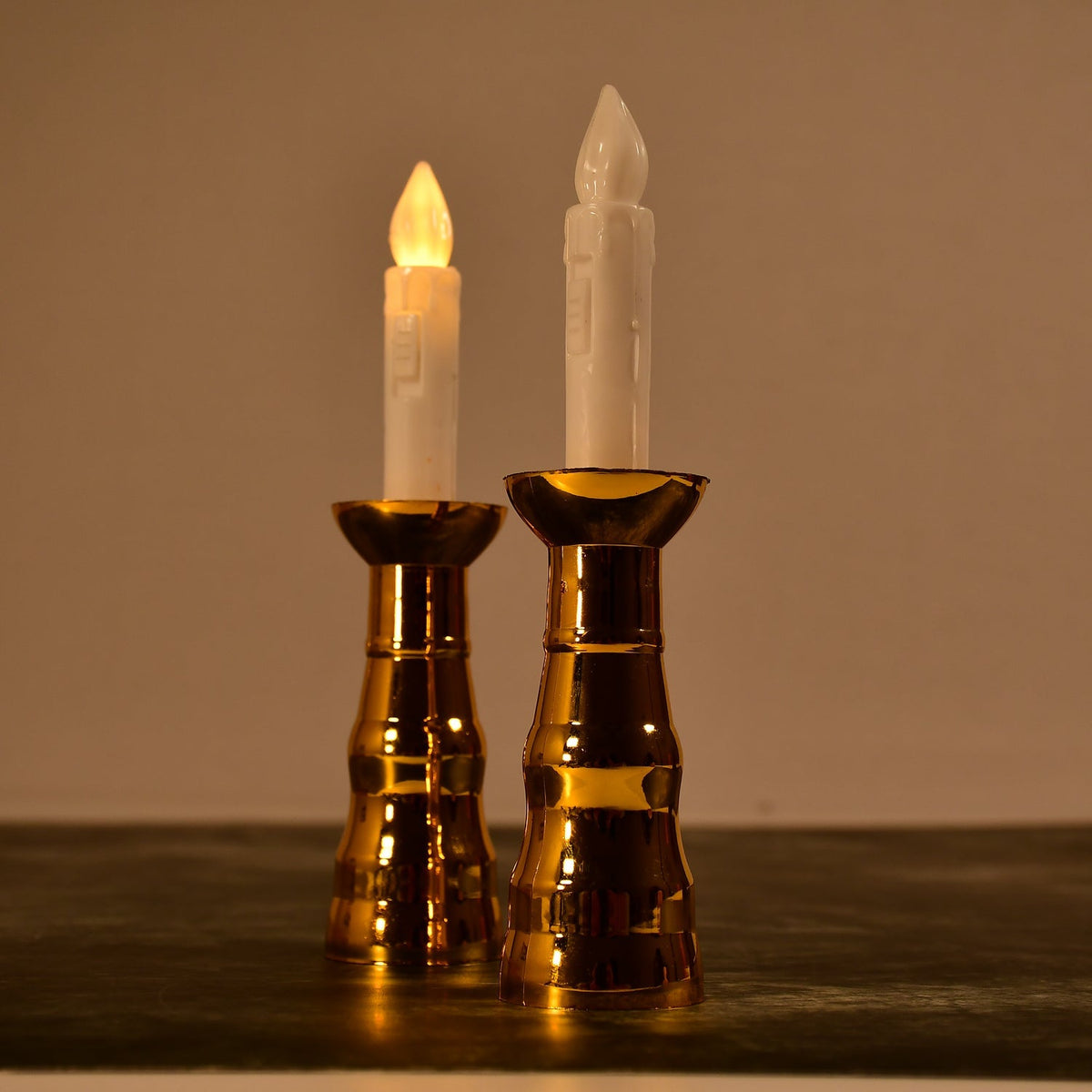 Two gold-based LED candles with illuminated flames stand on a dark surface in a dimly lit room.