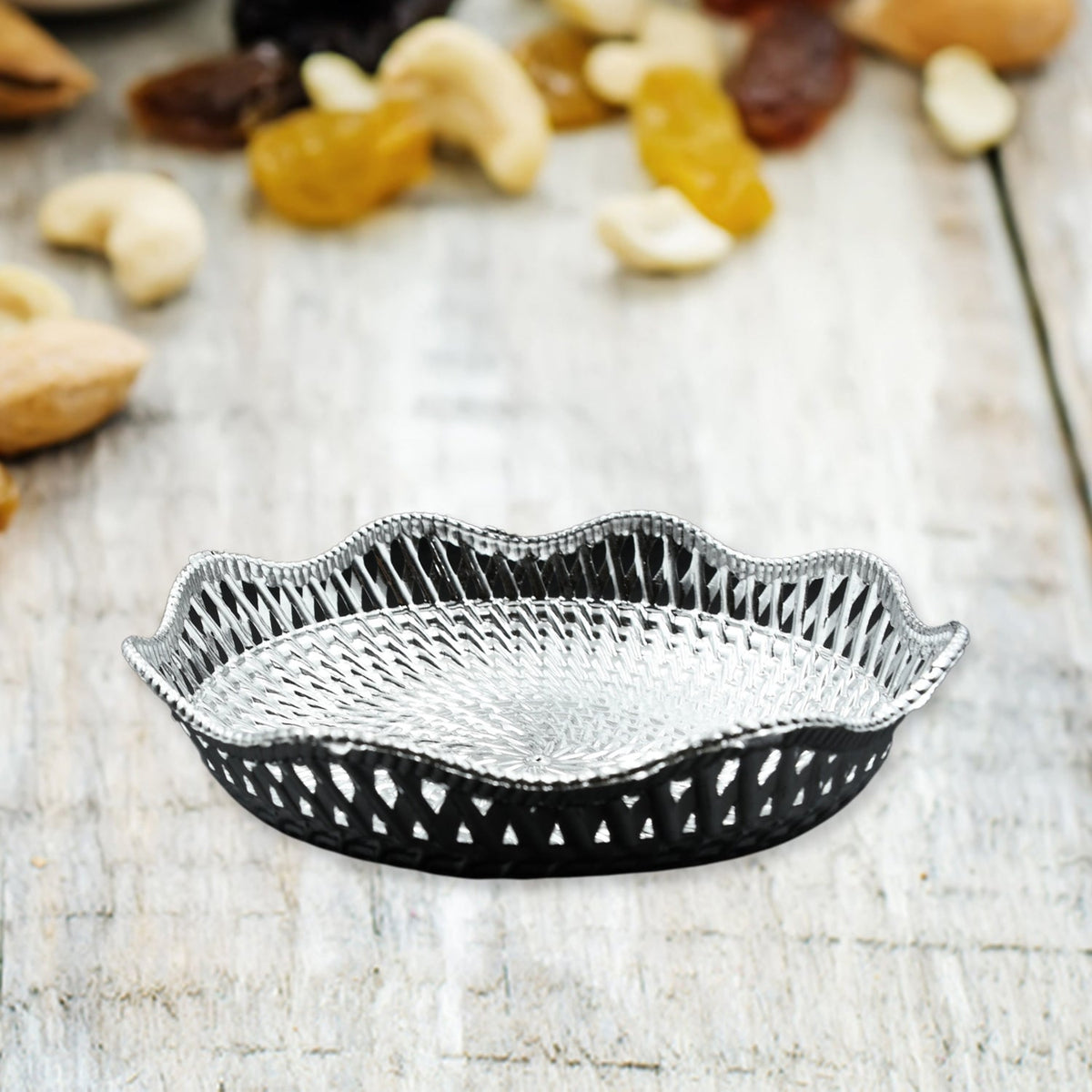 A silver plastic scalloped serving bowl with a textured bottom sits on a light wooden surface, surrounded by various nuts
