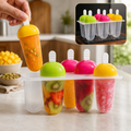 A hand holds an orange popsicle from a 6-piece mold with other fruit-filled popsicles, on a kitchen counter
