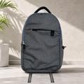 A grey backpack with a front zipper pocket and blue trim stands on a light grey surface next to a plant.