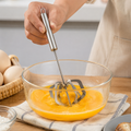 A person's hand holding a silver stainless steel hand whisk, mixing yellow liquid in a clear glass bowl.