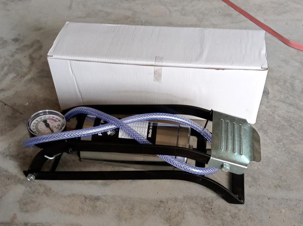 A black and silver foot pump with a hose and gauge is placed in front of a white cardboard box on a concrete floor.