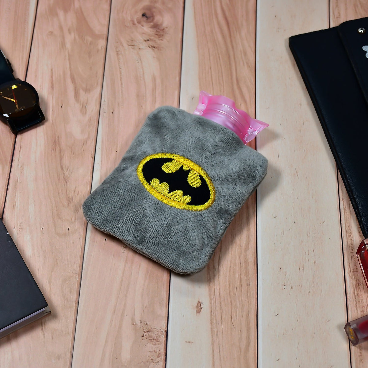 Gray plush hot water bag with a Batman logo on a wooden table with a watch and notebook.