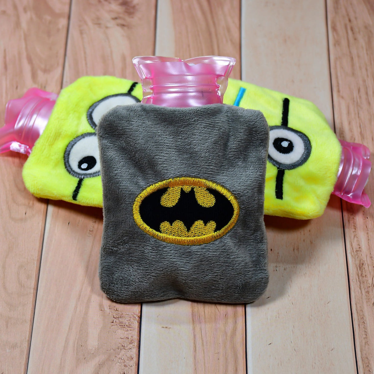 Gray plush hot water bag with Batman logo in front of a yellow Minion-themed hot water bag on wood.