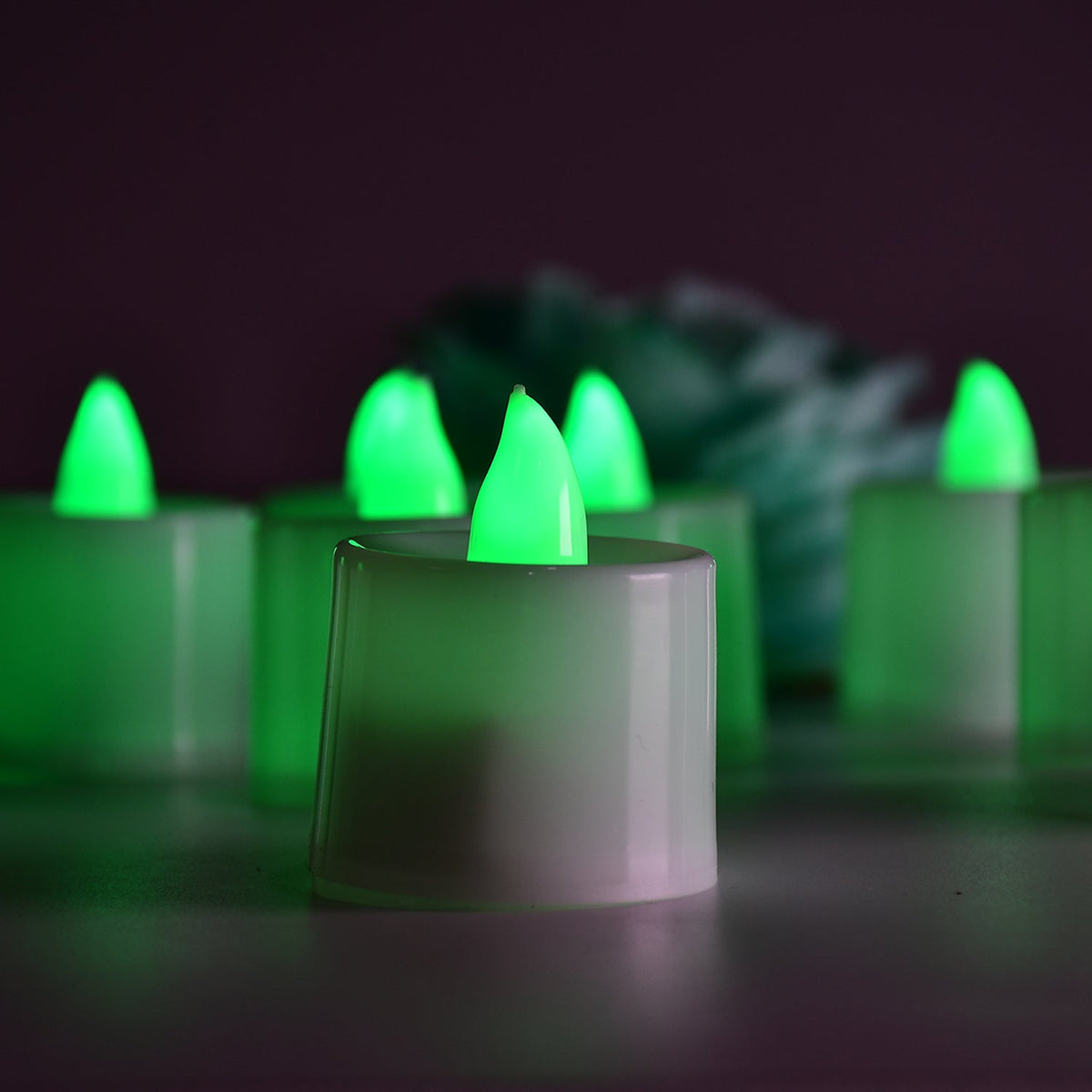 Six green flameless LED tealights glow in a dimly lit setting, with a soft teal flower in the blurred background.