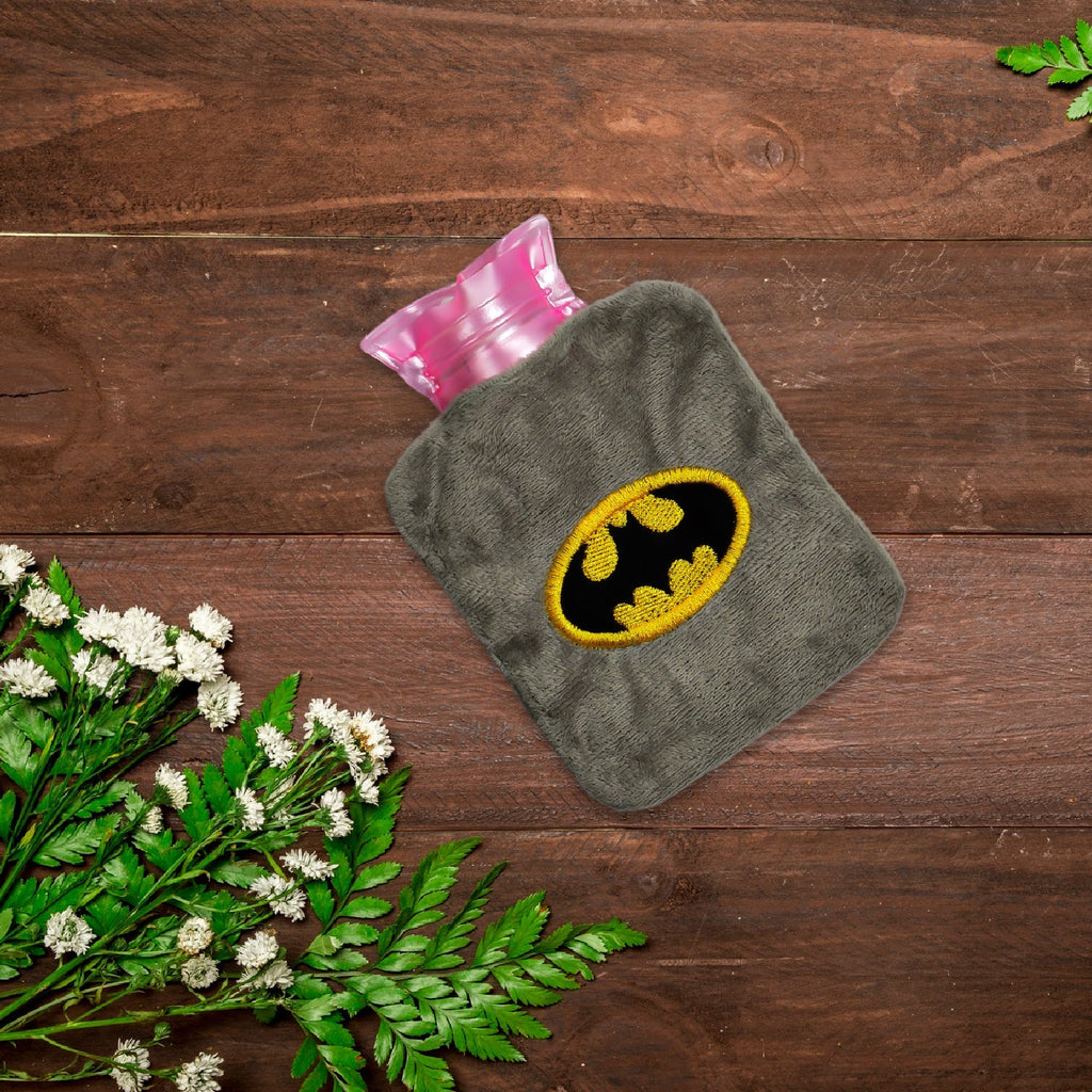 Gray plush hot water bag with a Batman logo on a dark wooden surface with white flowers and green leaves.