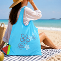 A woman sits on a beach towel with a light blue reusable shopping bag featuring red and white flower designs.