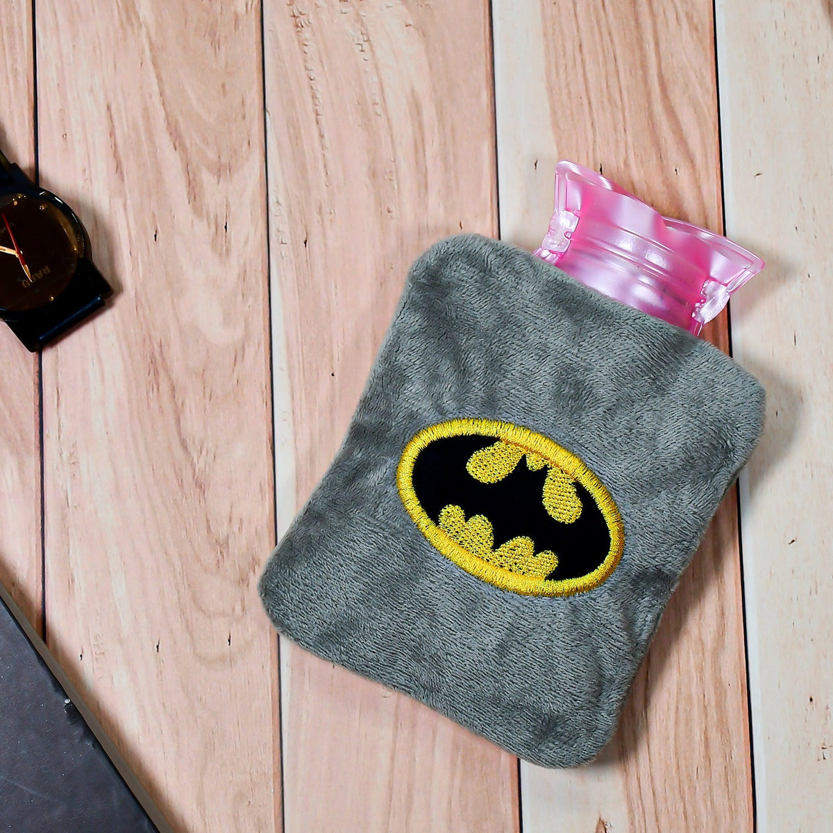Gray plush hot water bag with a Batman logo on a light wooden surface with a black watch and notebook.