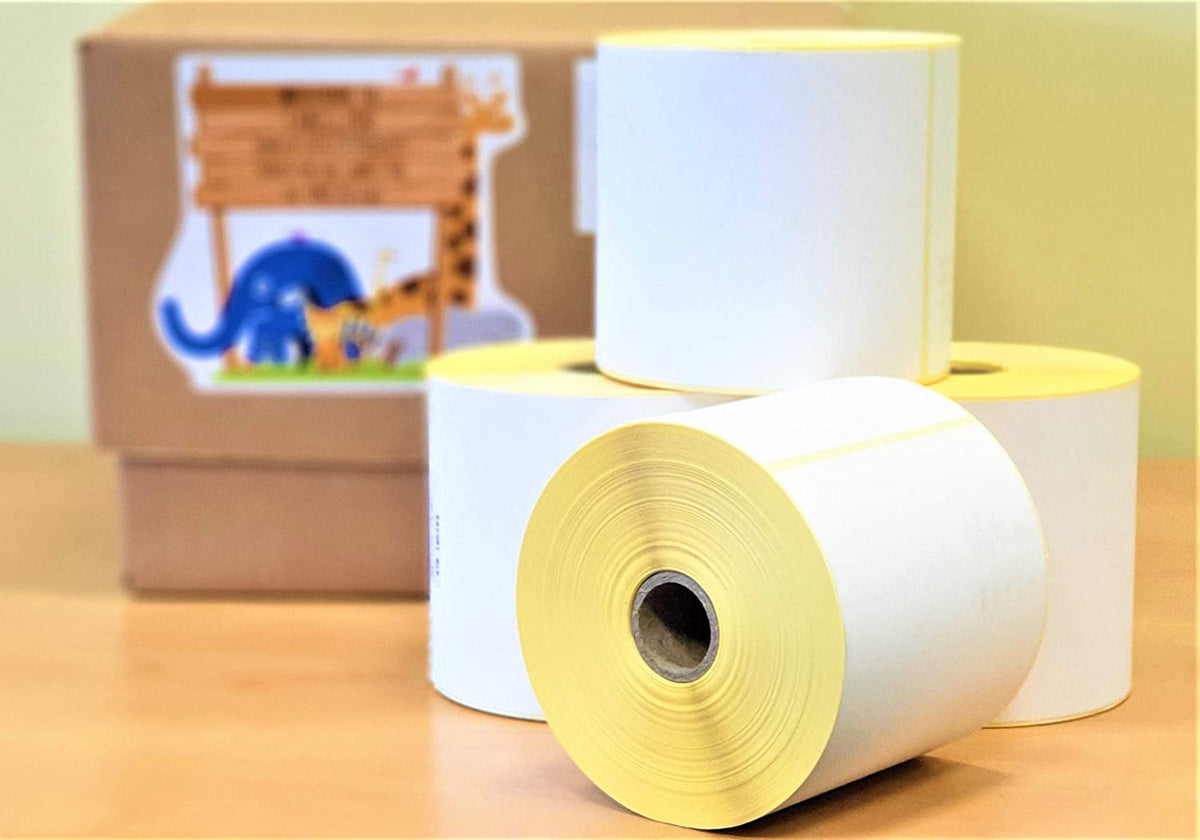 Four rolls of white thermal labels with yellow backing are stacked on a wooden surface, with a blurred brown box behind.