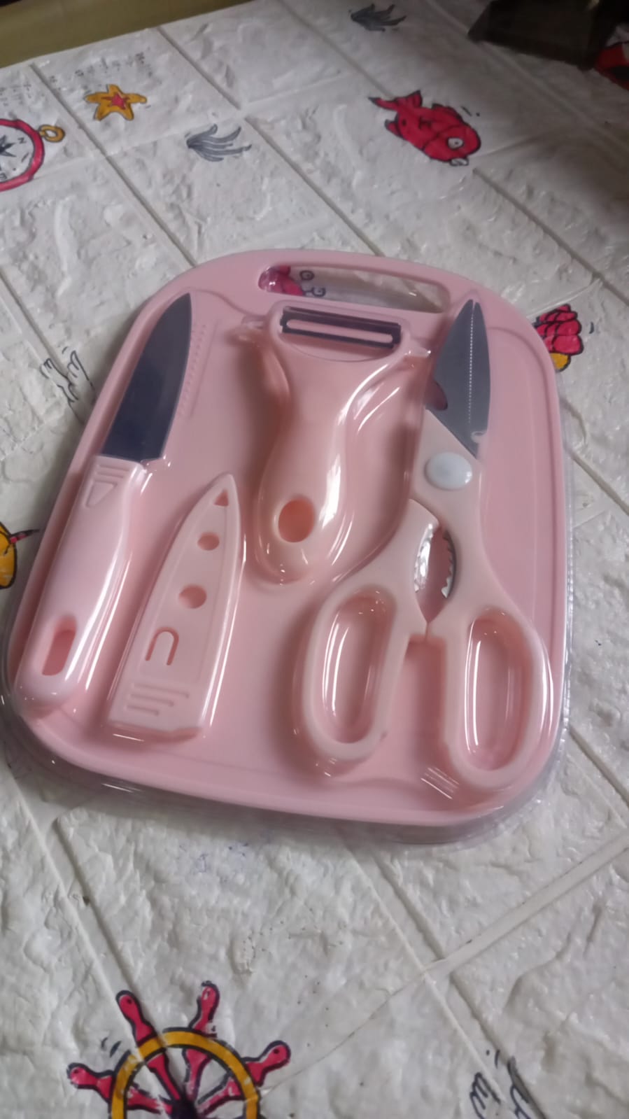 The pink cutting board set, including a knife, peeler, and scissors, is sealed in clear plastic packaging on a patterned