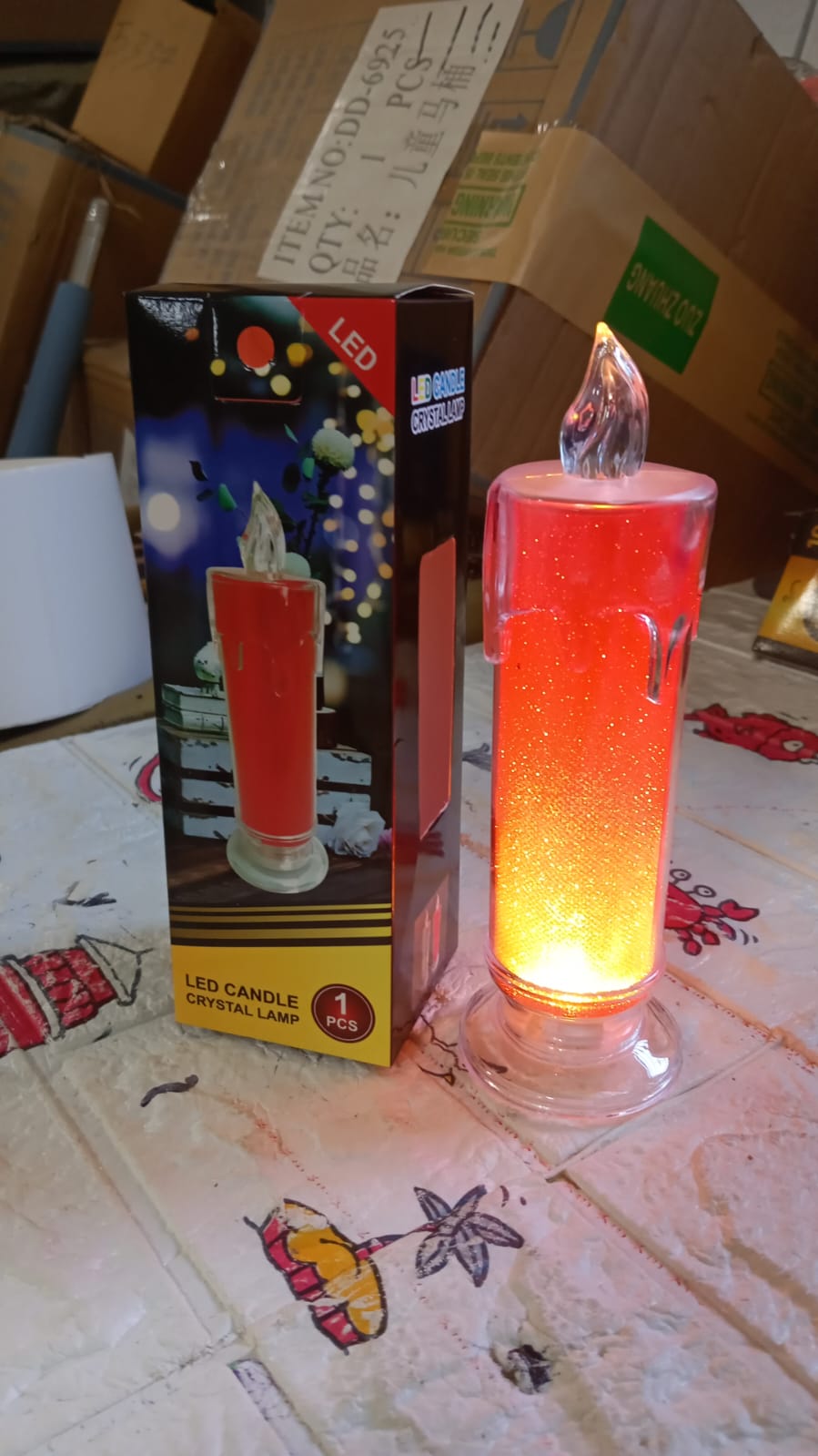A red LED glitter candle next to its packaging on a patterned surface, with a cardboard box in the background.