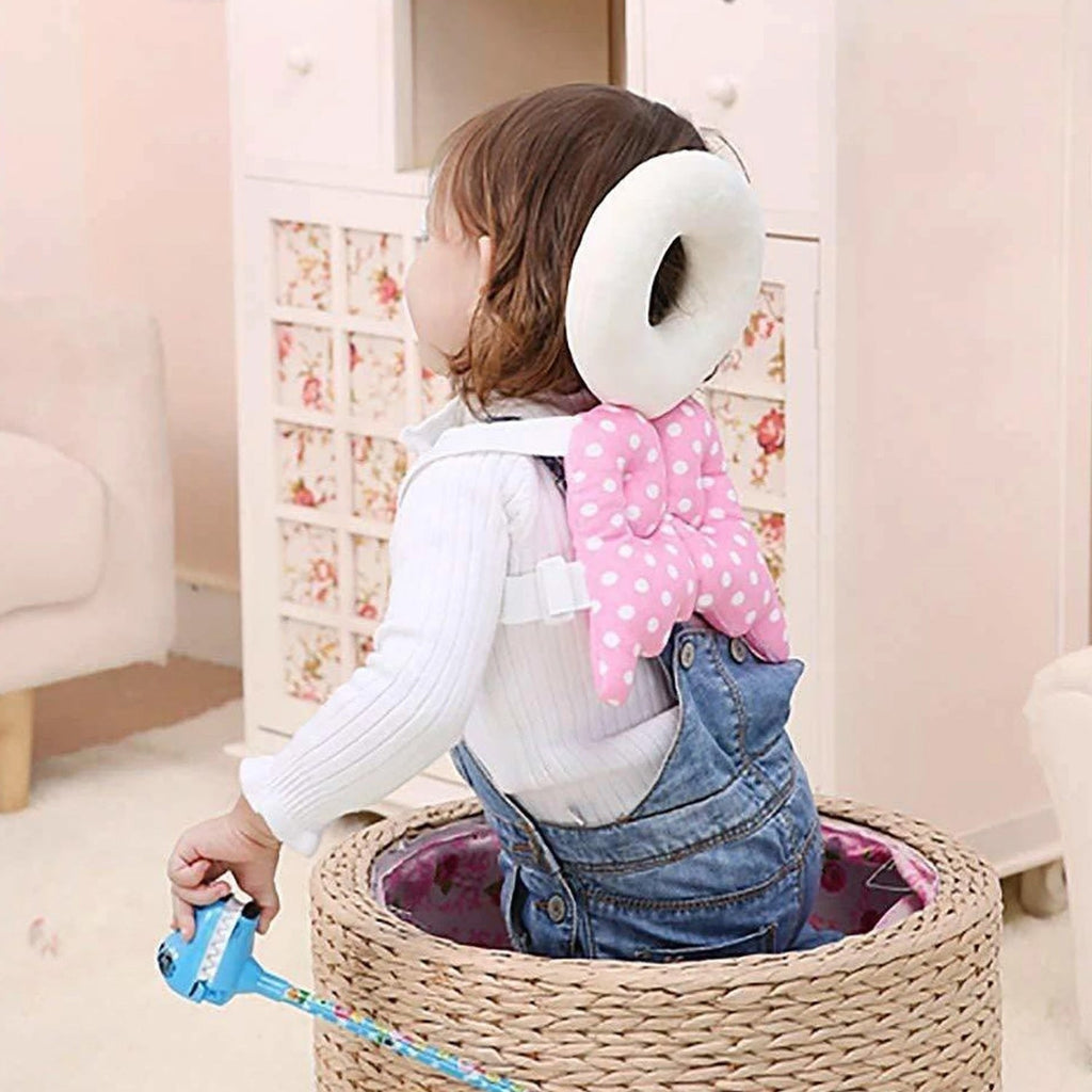 A toddler wearing a pink and white polka dot head protector with angel wings sits in a woven basket, looking over their