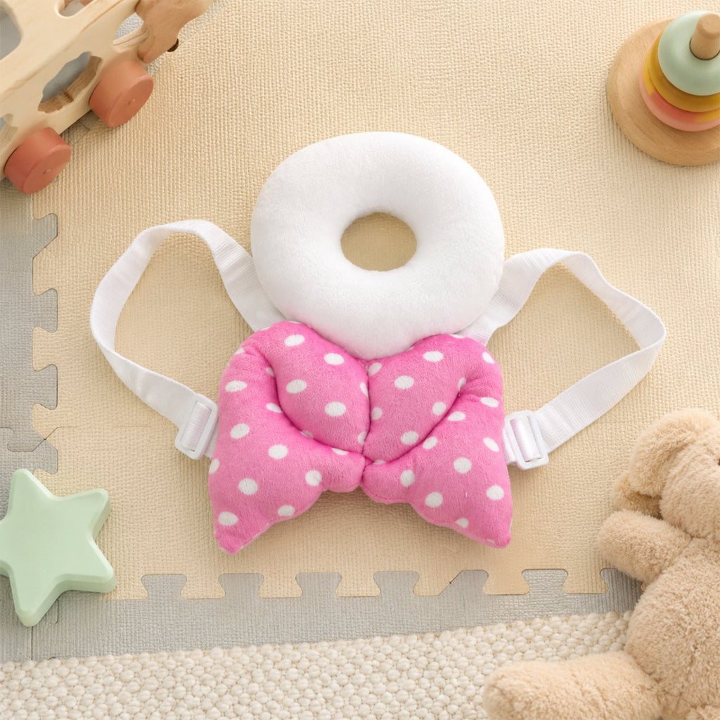 A pink and white polka dot head protector with angel wings lies flat on a beige playmat surrounded by toys.