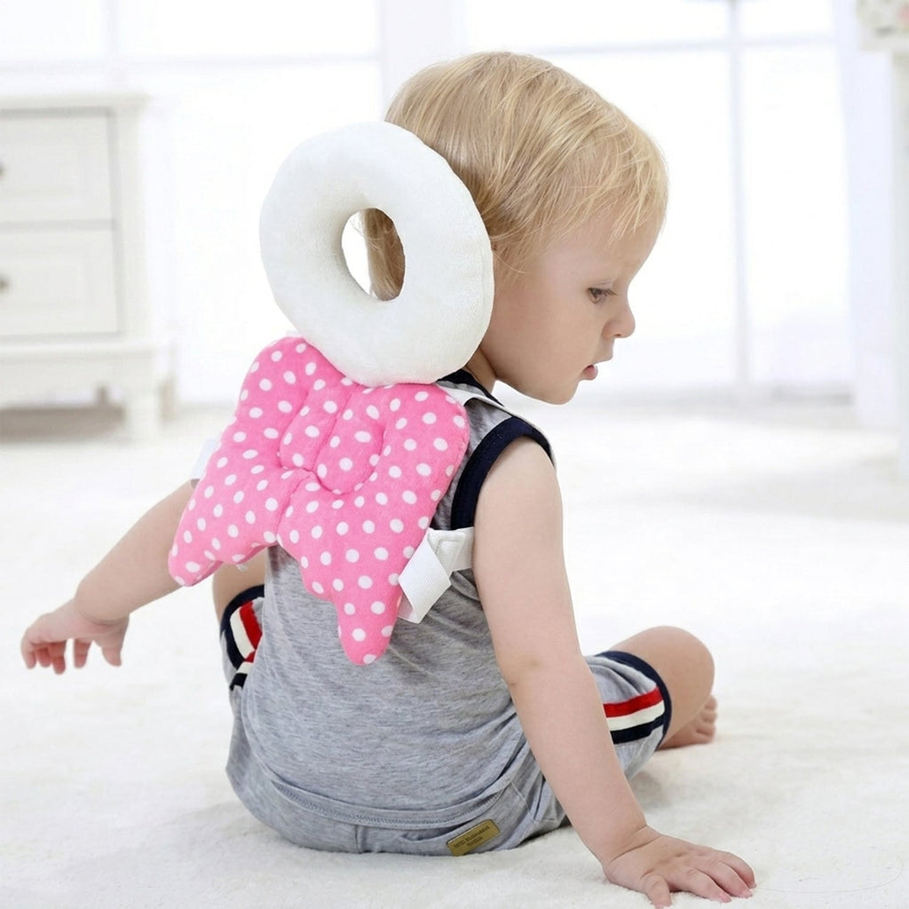 A toddler wearing a pink and white polka dot head protector with angel wings sits on a white rug looking to the side.