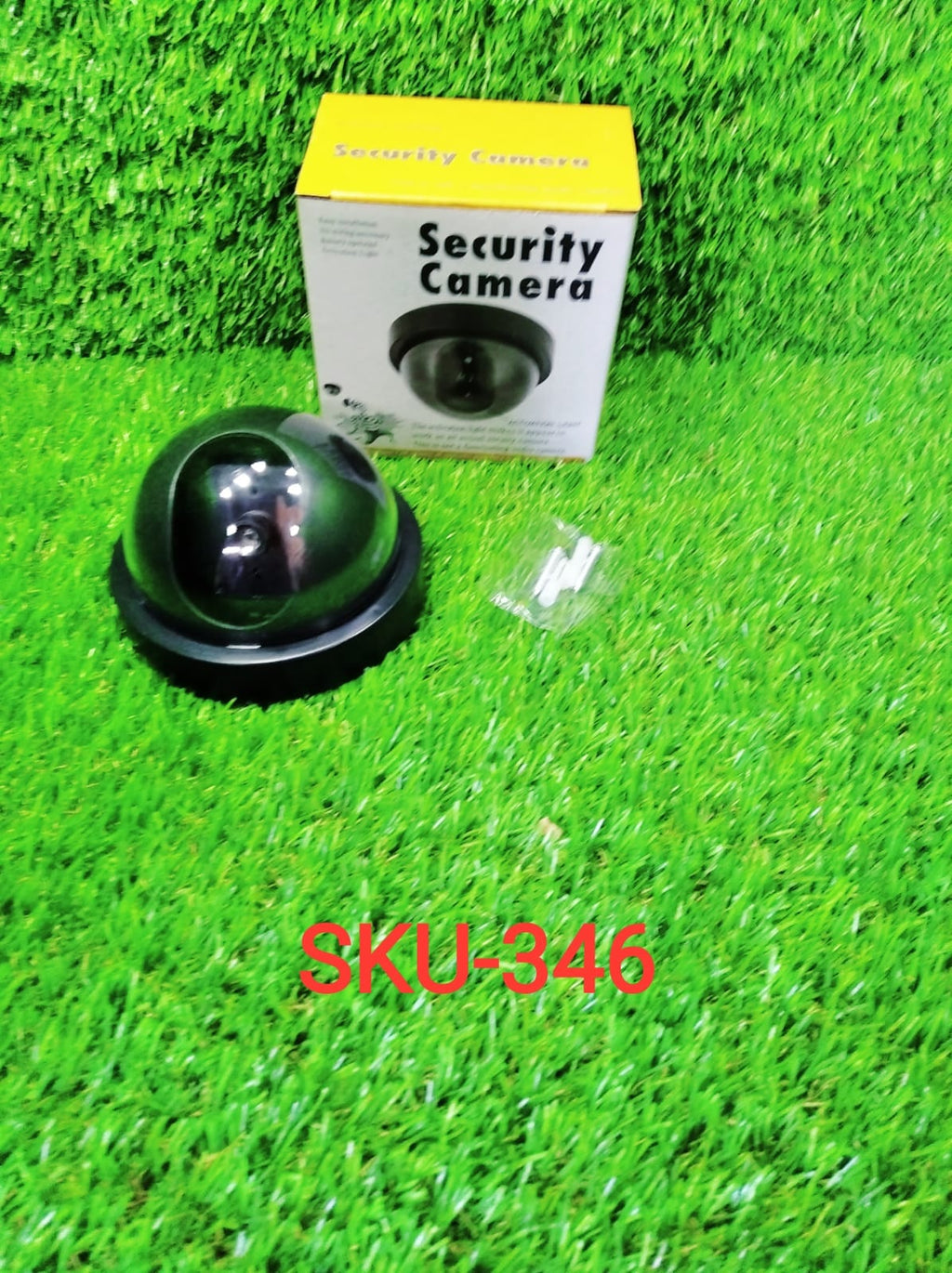 Black dome dummy security camera next to its yellow and white packaging box and small white screws, all on artificial green