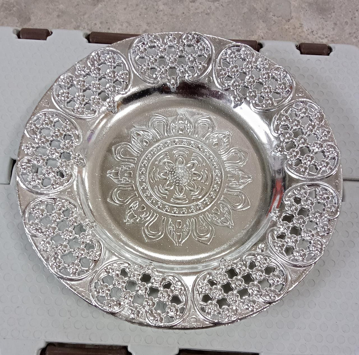 A silver-colored plastic pooja thali with a detailed embossed pattern resting on a light gray, textured surface.